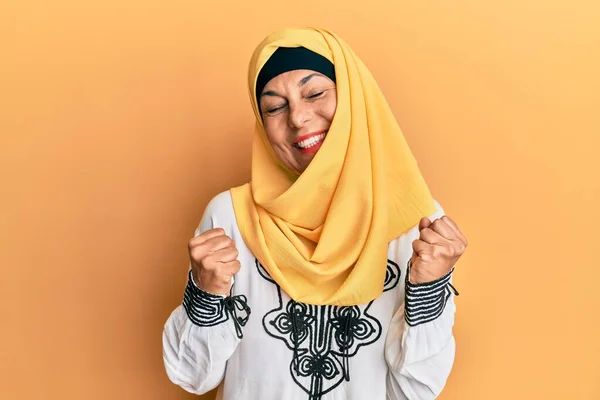 腕を上げて成功に興奮し 目を閉じて勝利笑顔を祝う伝統的なイスラムヒジャーブスカーフを身に着けている中世のヒスパニック女性 勝者のコンセプト — ストック写真