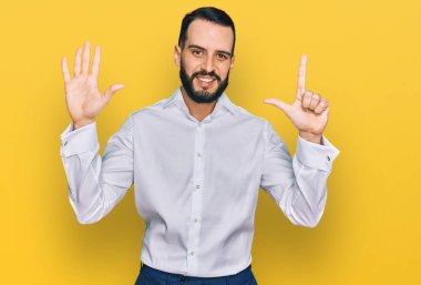 Sakallı genç adam iş gömleği giyiyor ve yedinci parmaklarıyla işaret ederken kendinden emin ve mutlu bir şekilde gülümsüyor.. 