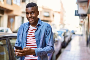 Genç Afrikalı Amerikalı adam şehirde akıllı telefon kullanarak mutlu bir şekilde gülümsüyor..