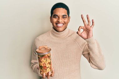 Genç, yakışıklı İspanyol adam elinde farfalle makarna kavanozuyla tamam işareti yapıyor, gülümsüyor ve mükemmel bir sembol oluşturuyor. 