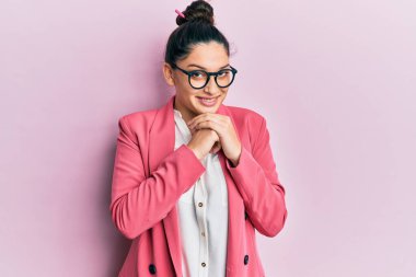 Orta Doğulu güzel bir kadın iş ceketi ve gözlük takıyor. Gergin ve heyecanlı bir şekilde gülüyor. Elleri çenesinde yan tarafa bakıyor. 