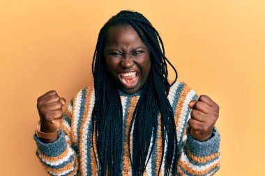 Örgülü genç siyahi kadın, öfkeyle bağırırken kızgın ve kızgın bir şekilde kazak giyiyor ve yumruklarını kaldırıyor. öfke ve agresif konsept. 