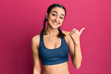 Genç esmer kız spor giysisi giyiyor ve örgüler örüyor. Gülümsüyor, gülümsüyor ve başparmağıyla yan tarafa bakıyor.. 