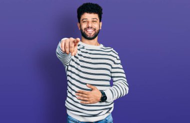 Sakallı, sıradan çizgili süveter giyen genç bir Arap sana gülüyor, eli vücudunun üzerinde kamerayı işaret ediyor, utanç ifadesi var. 