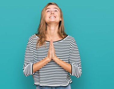 Güzel sarışın kadın gündelik giysiler giyip elleri bağlı yalvarıyor ve dua ediyor. Yüzünde umut ifadesi var. Duygusal ve endişeli. Yalvarıyor. 