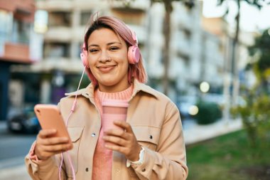 Genç İspanyol kız şehirde akıllı telefon ve kulaklık kullanarak kahve içiyor..
