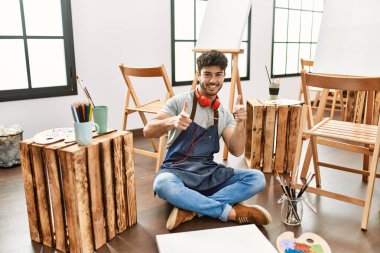 Sanat stüdyosunda oturan genç İspanyol adam mutlu ve komik yüzüyle kamerayı işaret ediyor. iyi enerji ve titreşimler. 