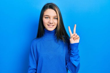 Balıkçı yaka kazak giyen genç esmer kız parmaklarını iki numara ile gösteriyor ve gülümsüyor. Kendinden emin ve mutlu.. 