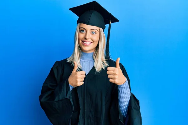 Beautiful blonde woman wearing graduation cap and ceremony robe success sign doing positive gesture with hand, thumbs up smiling and happy. cheerful expression and winner gesture.