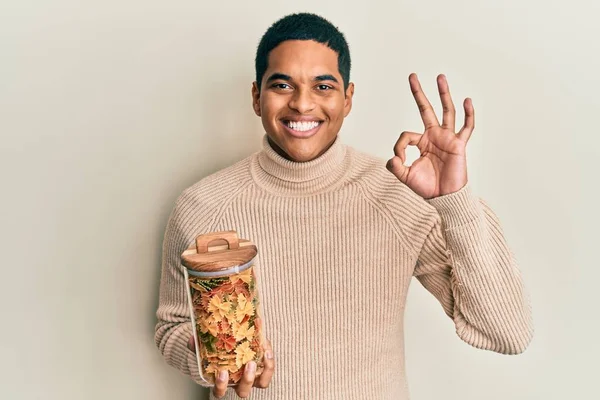 Genç, yakışıklı İspanyol adam elinde farfalle makarna kavanozuyla tamam işareti yapıyor, gülümsüyor ve mükemmel bir sembol oluşturuyor. 