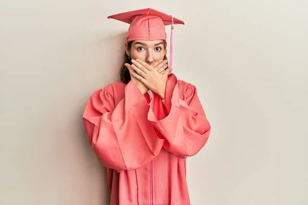 卒業キャップと儀式用のローブを着た若い白人女性がミスのために手で口を覆いショックを受けました 秘密の概念 — ストック写真