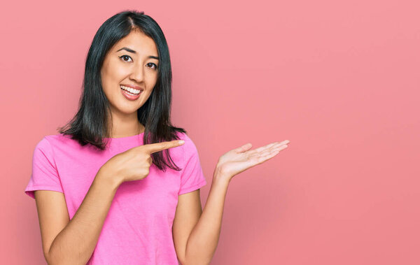 Beautiful asian young woman wearing casual pink t shirt amazed and smiling to the camera while presenting with hand and pointing with finger. 