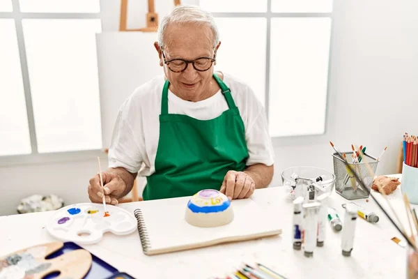在艺术工作室的桌子上坐着一位头发灰白的资深画家 他高兴地微笑着 画着陶器 — 图库照片