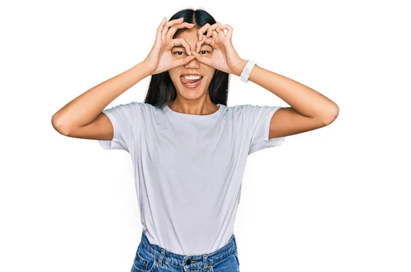 漂亮的年轻亚洲女人穿着休闲的白色T恤 像双筒望远镜一样做手势 伸出舌头 眼睛透过手指看着 疯狂的表达 — 图库照片