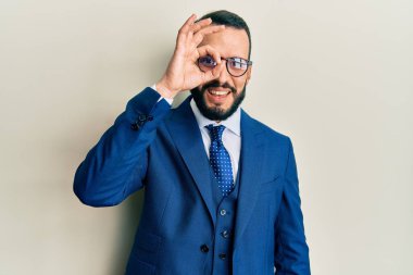 Sakallı, takım elbiseli ve kravatlı genç adam el hareketleriyle gülümsüyor ve mutlu yüzüyle parmaklarının arasından bakıyor.. 