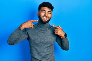 Balıkçı yaka kazak giyen, gülümseyen, parmaklarıyla ve ağzıyla işaret eden sakallı bir Arap. diş sağlığı kavramı. 