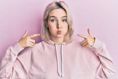 Genç sarışın kız dudaklarını parmaklarıyla göstererek komik yüzlü yanaklarını şişiriyor. Ağzı havayla şişer, havayı yakalar.. 