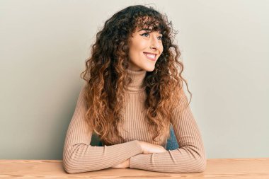 Latin asıllı genç bir kız, gündelik giysiler içinde masada oturuyor, yüzünde gülümsemeyle yan yana bakıyor, doğal bir ifade. Kendinden emin gülen. 