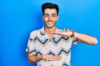 Latin asıllı genç adam, sıradan kıyafetler giyiyor. Elleriyle işaret ediyor. Ölçü sembolü, büyük boy işareti. Kameraya bakıp gülümsüyor. ölçüm kavramı. 