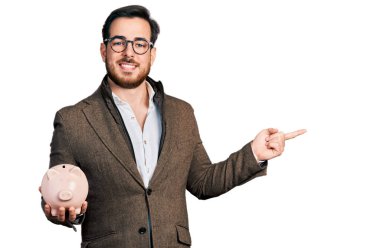 Genç İspanyol adam domuzcuk kumbarasını tutuyor gülümsüyor mutlu bir şekilde eli ve parmağı yana doğru işaret ediyor 