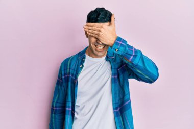 Yakışıklı, sıradan kıyafetler giyen, gülümseyen ve elleri yüzünde, şaşırtmak için gözlerini kapatan İspanyol bir adam. kör kavram. 