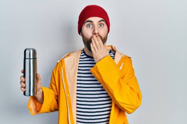 Sakallı, termosu olan beyaz bir adam ağızını eliyle kapatıyor, şokta ve hata yapmaktan korkuyor. şaşırmış ifade 