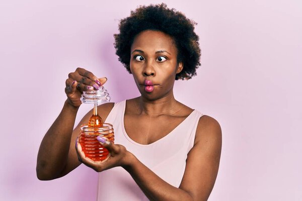 Young african american woman holding honey making fish face with mouth and squinting eyes, crazy and comical.