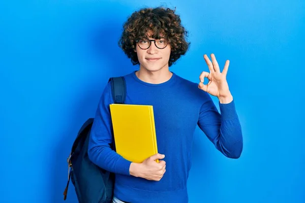 Handsome young man wearing student backpack and holding book doing ok sign with fingers, smiling friendly gesturing excellent symbol
