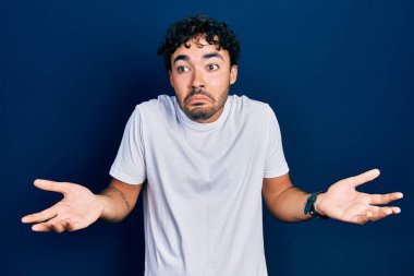 Gündelik beyaz tişört giyen, kolları ve elleri havada, kafası karışık, genç İspanyol bir adam. şüphe kavramı. 