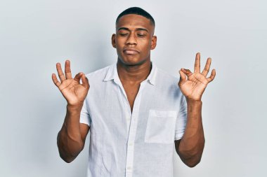 Genç siyah adam, günlük beyaz gömlekler giyip rahatlıyor ve gözleri kapalı gülümsüyor parmaklarıyla meditasyon hareketi yapıyor. yoga konsepti. 