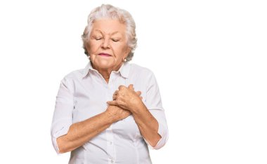 Üstün gri saçlı, günlük kıyafetler giyen, elleri göğsünde, gözleri kapalı gülümseyen ve yüzünde minnet ifadesi olan bir kadın. sağlık kavramı. 