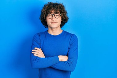 Sıradan kıyafetler ve gözlük takan yakışıklı genç adam kameraya bakarken kollarını kavuşturup gülümsüyor. pozitif kişi. 