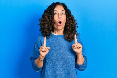 Orta yaşlı İspanyol kadın gündelik kıyafetler giyip şaşkın şaşkın yukarı bakıyor ve parmaklarıyla işaret ediyor ve kollarını kaldırıyor.. 