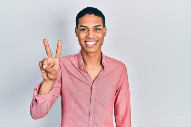 Genç Afro-Amerikan adam, günlük kıyafetler giyip kameraya gülümseyen ve zafer işareti yapan. İki numara.. 