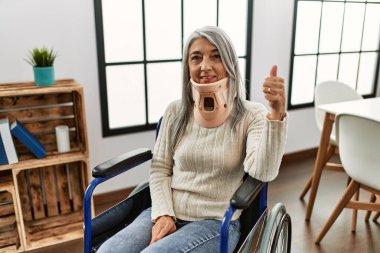 Orta yaşlı, gri saçlı, tekerlekli sandalyede oturan, tasmalı, mutlu ve mutlu bir şekilde gülümseyen, başparmağıyla mükemmel ve onay işareti yapan bir kadın. 