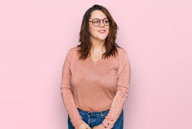 Genç ve cüsseli bir kadın günlük kıyafetler ve gözlük takıyor gülümsüyor, yan tarafa bakıyor ve düşünüyor.. 