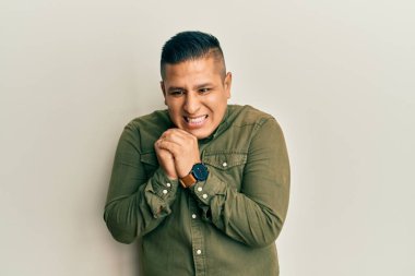 Gündelik kıyafetler giyen genç latin adam gergin ve heyecanlı, elleri çenesinde yan tarafa bakıyor. 