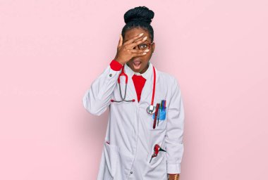 Genç Afro-Amerikan kadın doktor üniforması giyiyor ve steteskop ile yüzünü ve gözlerini kapayarak utangaç bir ifadeyle parmaklarının arasından bakıyor.. 