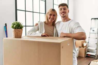 Genç beyaz çift mutlu bir şekilde gülümsüyor ve yeni evlerinde karton kutuya yaslanıyorlar..