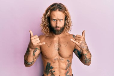 Sakallı, uzun saçlı, tişörtsüz, Shaka işareti yapan, şüpheci ve sinirli, sorun yüzünden surat asan yakışıklı bir adam. negatif kişi. 