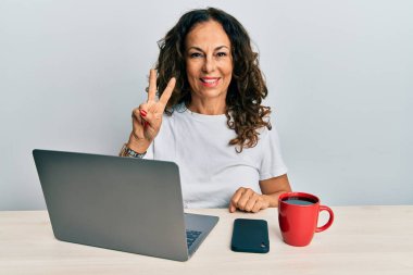 Ofiste çalışan orta yaşlı güzel bir kadın bilgisayarını kullanarak kameraya gülümseyen bir gülümsemeyle zafer işareti yapıyor. İki numara.. 