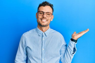 Gündelik kıyafetler ve gözlük takan genç İspanyol adam mutlu bir şekilde gülümsüyor ve avuç içi kameraya bakıyor.. 