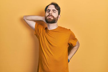 Sakallı, sıradan sarı tişörtlü, boynu ağrıyan, eliyle boynuna dokunan beyaz bir adam. 