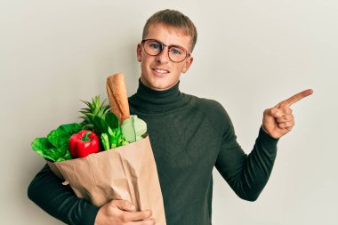 Elinde alışveriş poşeti olan beyaz bir genç adam gülümsüyor mutlu bir şekilde elini ve parmağını kenarı işaret ediyor. 
