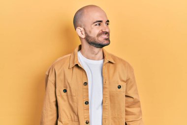 Gündelik kıyafetler giyen genç kel bir adam yüzünde gülümsemeyle yan yana bakıyor, doğal bir ifade. Kendinden emin gülen. 