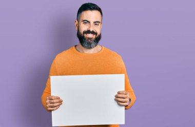 Sakallı, boş afiş tutan, yüzünde mutlu ve havalı bir gülümsemeyle gülümseyen İspanyol bir adam. diş gösterme. 