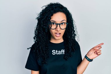 Kıvırcık saçlı, baston giyen genç İspanyol kadın. Tişörtü parmağıyla yana dönük, ağzı açık, şaşkın bir ifade sergiliyor.. 