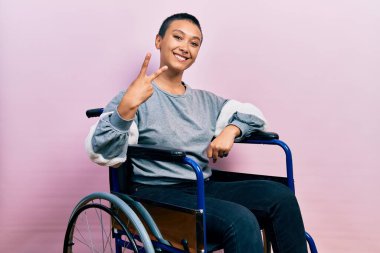 Kısa saçlı, ispanyol bir kadın tekerlekli sandalyede oturuyor ve iki numaralı parmaklarıyla işaret ediyor. Kendinden emin ve mutlu bir şekilde gülümsüyor.. 