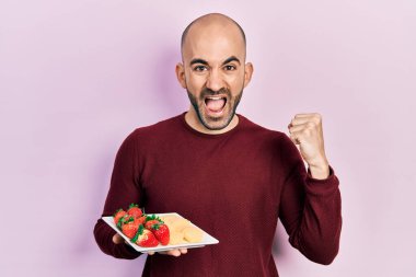 Genç kel adam taze ve sağlıklı meyveler yiyor gururla bağırıyor, zaferi kutluyor ve başarıyı kollarını kaldırarak kutluyor. 