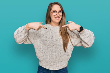 Gündelik süveter ve gözlük takan genç sarışın kadın. Kendine güvenen, gülümseyen, kendini gururlu ve mutlu parmaklarıyla gösteren.. 